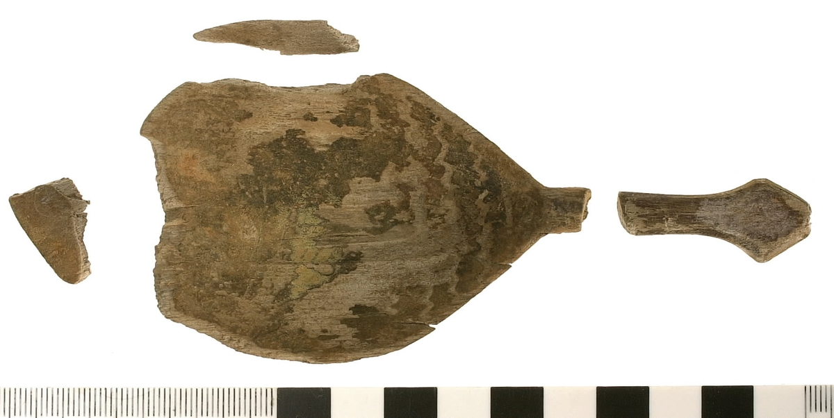 Fyra delar av en träsked. De två större delarna utgör skedskaftet samt större del av skedbladet med ett mindre parti av skaftet. De övriga två delarna utgör fragment av skedblad.

Insidan av skedbladet är dekorerad med ett ristat/glödritat vågmönster. Skedskaftet är kort och rakt, med profilerad skaftände. Längst ut på skaftänden syns en brottyta. Troligtvis har skaftet avlutats i en ändknopp. Skeden har gul-röda utfällningar samt rödbruna färgspår.

Skeddelarna är mycket ömtåliga, med sprickor.

För mer information se Dokumentationsblankett under Referenser – Länkade filer.