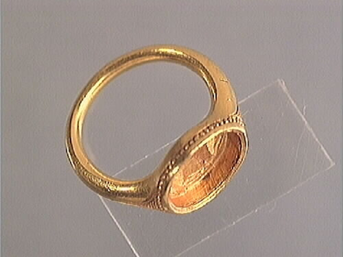 En fingerring av guld.
Guldringen har en oval infattning. Runt infattningen löper en pärlliknande bård. Bården består av 60 små och pärlliknande guldkulor, om 1 mm i diameter. På var sida om infattningen sitter en ingraverad, avsmalnande triangel försedd med horisontella streck. Ringen har varit försedd med en sten eller sigill vilken nu saknas. Ringens vikt uppgår till 10 gram.
Ringen är i mycket gott skick.

Text in English: Robust finger ring of gold with oval setting. The stone is missing.

Ingraved round the setting is a pearl-like border consisting of 60 tiny circles, 1 mm in diameter, and on either side of the setting - on the ring''s ''shoulders'' - is an inward curved triangle with horizontal lines.

Weight: 10 gram.
In very good condition.