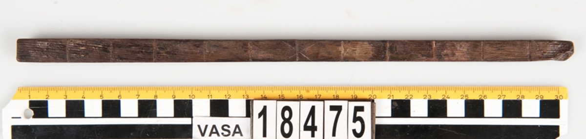 En tumstock av trä.

Tumstocken är försedd med elva inristade skåror som markerar tolv tum. På den mittersta skåran finns ett inristat kryss. Ytterligare ett kryss, dock något mindre, har ristats in vid skåran närmast mittskåran.

Tumstocken är i gott skick.

För mer information se Dokumentationsblankett under Referenser – Länkade filer.