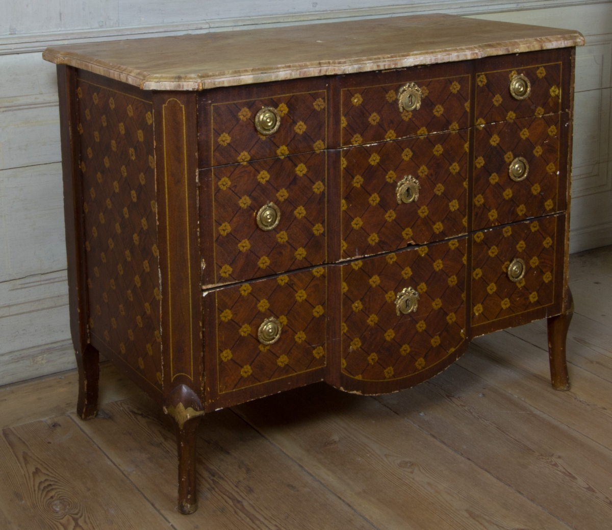 Byrå av furu, med imitationsmåleri som efterliknar fanér och inläggningar i olika träslag, i form av rutinläggningar och blommor. Med marmorerad skiva av trä. Rak form med avfasade hörn, med svagt svängda ben. Fronten med förkroppat mittparti, som nedåt avslutas i svagt bågformat undergehäng. Med tre lådor med nyckelbrickor med bandrosetter och beslag med dragringar i form av lagerkransar av förgylld mässing.