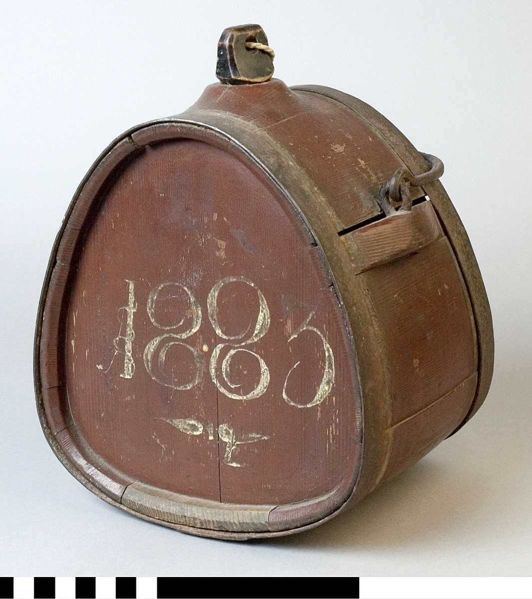 Mjölkflaska av trä, målad brun. Med band och handtag av järn. Med tillhörande propp av svartmålat trä. 
Märkt på ena sidan 1883, i vitt.
Märkt på andra sidan "JMS", i vitt.