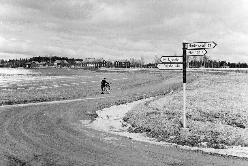 Lantbrevbärare, postiljon Erik Bergman (cykelåkande) i
Delsbo.  April 1956. Brevbäringsturen är 2 ½ mil lång och omfattar nästan 300 hushåll.  Postväskan väger ofta 30-40 kg.