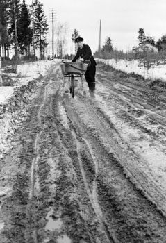 Lantbrevbärare, postiljon Erik Bergman (cykelåkande) i
Delsbo.  April 1956.

Brevbäringsturen är 2 ½ mil lång och omfattar nästan 300 hushåll. Postväskan väger ofta 30-40 kg.  På de lerigaste och värsta sträckorna måste cykeln ledas.