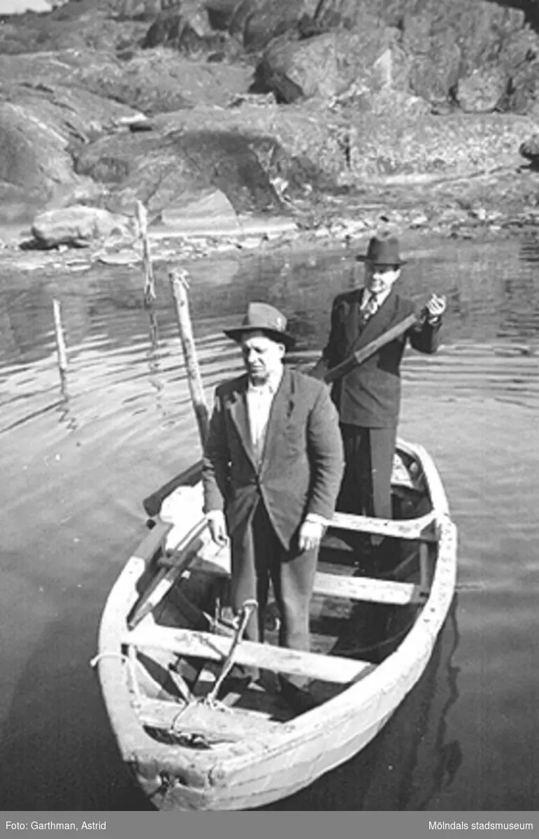 Från vänster: Helmer Garthman och Dimitri Korski står i en eka som ligger i vattnet. De är både fiske- och arbetskamrater. Klippor syns i bakgrunden vid Näset, 1960-tal.