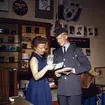 Brevbärare iklädd postens uniform från 1962 års beklädnadsreglemente, överlämnar post till kvinnlig expedit i en fotobutik.