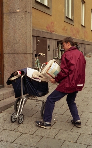 Brevbärare i Stockholms innerstad, Polhemsgatan. Vinröd jacka och
mörkblå byxor. Stor postkärra försedd med handbroms. Stor mängd
reklam.