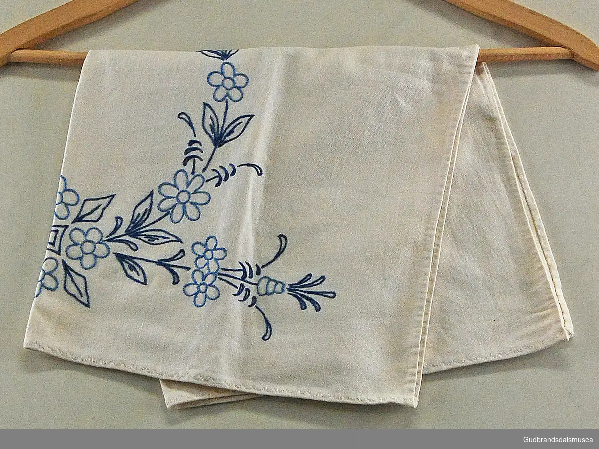Duk, tilnærmet kvadratisk brikke i hvitt/beige, med brodert blomster- og blad-mønster i ring. Motiv brodert i lys- og mørkblå tråd. 