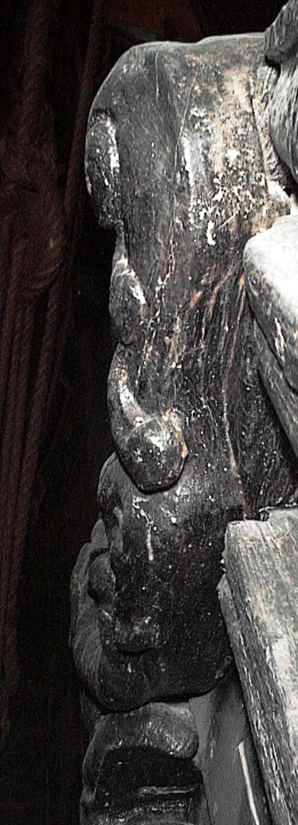 Skulptur av en triton, svängd mot höger.
Tritonen är framställd i vänster profil och avtecknar sig i hög relief mot det släta bakstycket. Tritonen har en stor näsa och treflikig mustasch. På huvudet sitter en hjälm med dekorativa bladvoluter. Armarna är ersatta av långa, flikiga bladslingor. Bålens övergång i den fjällförsedda fiskstjärten markeras av stora bladbildningar. Tritonens stjärt döljs bakom stjärten och vingen till en under liggande drake, se fyndnummer 21277. På baksidan finns urtag för relingslisten.
Skulpturen är mycket välbevarad.

Text in English: A sculpture of a Triton curved to the right.
The figure is seen in left profile, in high relief against a plain back board. The face has a large nose and a moustache that splits into three parts. On its head is a helmet with decorative leaf volutes. The arms are replaced by long, lobed leaf formations. The transition area between the male torso and the scaled fish-tail is decorated with large leaves. The rear is hidden behind a dragon''s coiled tail and broad wing situated below, see No. 21277. There are notches on the back that fit into the railing.
The sculpture is very well preserved.