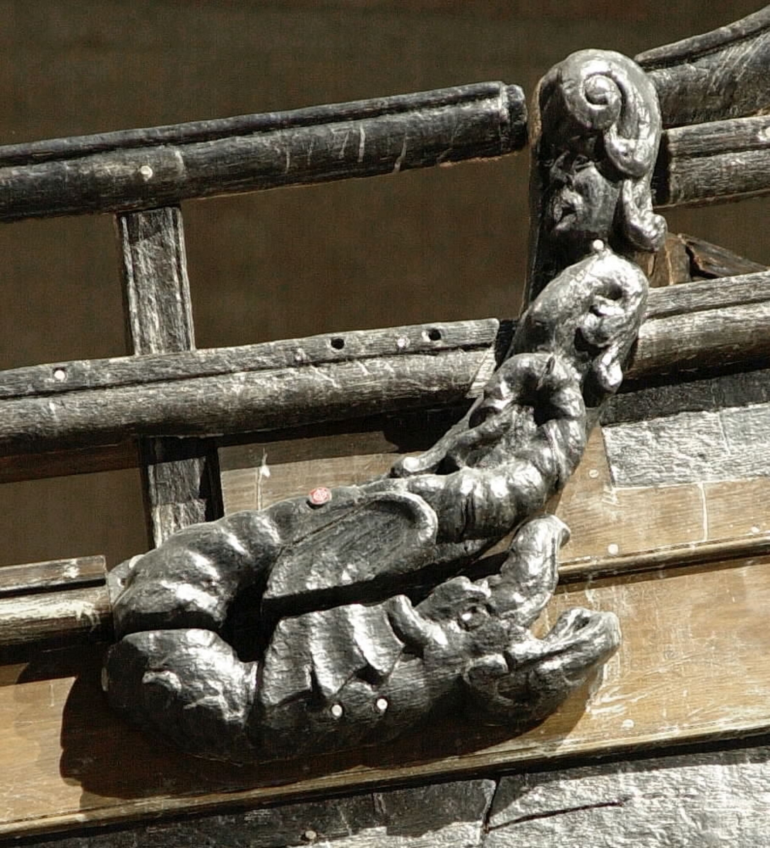 Skulptur av en triton, svängd mot höger.
Tritonen är framställd i vänster profil och avtecknar sig i hög relief mot det släta bakstycket. Tritonen har en stor näsa och treflikig mustasch. På huvudet sitter en hjälm med dekorativa bladvoluter. Armarna är ersatta av långa, flikiga bladslingor. Bålens övergång i den fjällförsedda fiskstjärten markeras av stora bladbildningar. Tritonens stjärt döljs bakom stjärten och vingen till en under liggande drake, se fyndnummer 21277. På baksidan finns urtag för relingslisten.
Skulpturen är mycket välbevarad.

Text in English: A sculpture of a Triton curved to the right.
The figure is seen in left profile, in high relief against a plain back board. The face has a large nose and a moustache that splits into three parts. On its head is a helmet with decorative leaf volutes. The arms are replaced by long, lobed leaf formations. The transition area between the male torso and the scaled fish-tail is decorated with large leaves. The rear is hidden behind a dragon''s coiled tail and broad wing situated below, see No. 21277. There are notches on the back that fit into the railing.
The sculpture is very well preserved.