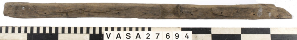 Relingslist. Den ena kortänden är uppfläkt. Den andra kortänden avslutas med en snedfasning, sannolikt en lask. Den andra slutar tvärt i sambands med en ursparning (sannolikt platsen för en årtull). Tolv stycken ej genomgående spikar i vad som sannolikt är den sida som vetter mot bordläggningen indikerar att den har spikats fast utifrån. Ett kraftigt genomgående hål syns i anslutning till ursparningarna för årtullarna. Spikhål syns i dessa avtrycks hörnor. På den sidan som vänder mot bordsidan och på undersidan syns snedfasade urtag, sannolikt för topparna till intimmer. I dessa urtagningar kan man skönja tydliga verktygsspår.