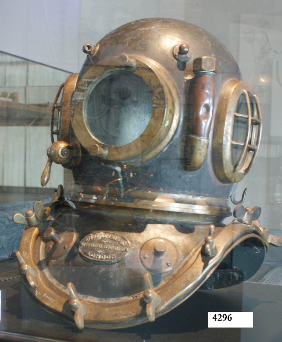 Dykarhjälm med bröstplåt. Material: koppar, mässing och glas. Från år 1880. Märkning: På bröstplåten: "Siebe, Gorman och Co Submarine Egineers London". På bröstplåtens skenor: "Siebe Gorman o Co".
