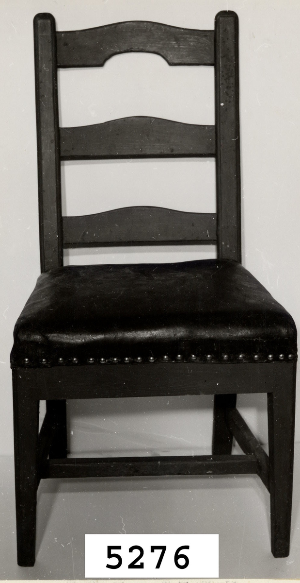 Stol av björk, rödmålad. Fyrsidiga ben med tre slåar, sk. H-kryss. Stoppad sits klädd med ryssläder. Tre tvärslåar i ryggstödet, den översta med urtag i nederkant.