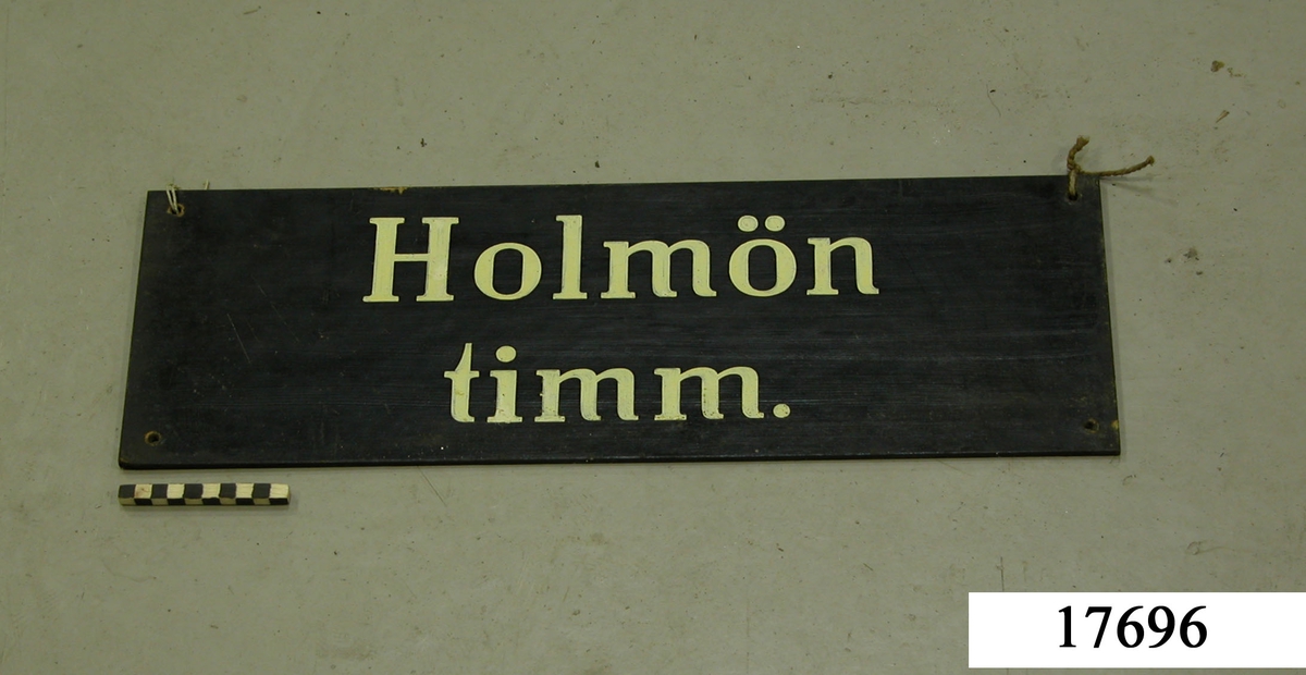 Rektangulär skylt av masonit, framsidan svartmålad, hål för upphängning. Vit schablonmålad text: " Holmön timm.".