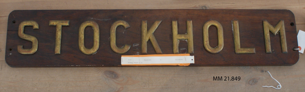 Namnbräda av teak, fernissad. Bokstäver av mässing: Stockholm.
X 136