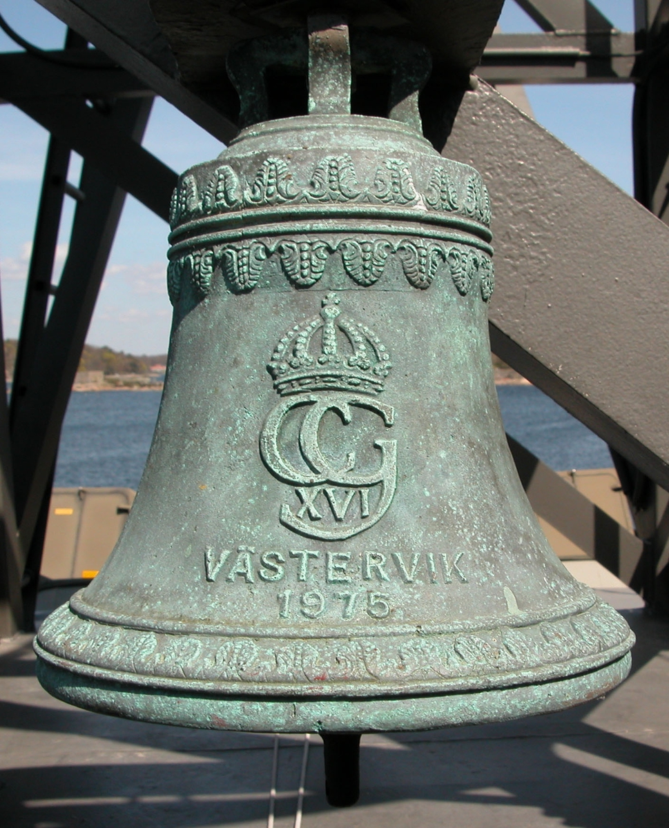 HMS Västeviks skeppsklocka av malm med kläpp. Grönärgad. På klockan finns Carl XVI Gustavs namnschiffer och texten "VÄSTERVIK, 1975".