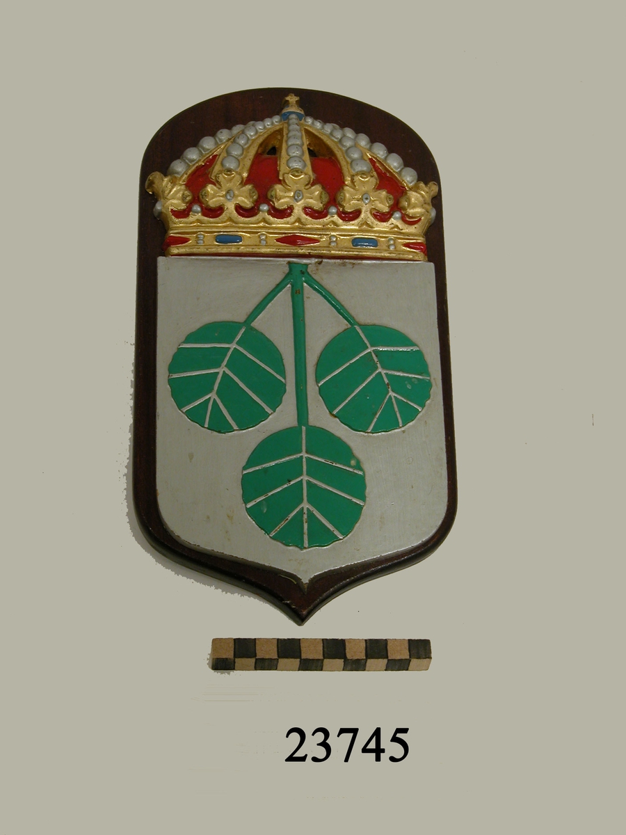 Vapensköld, minsvepare Aspö. Krönt vapensköld av gjuten mässing monterad på fernissad platta av trä. Bottenfärg i silver försedd med tre stiliserade, gröna aspblad som utgår från sköldens överkant. Bladnerver i silver. På baksidan liten krok för upphängning.