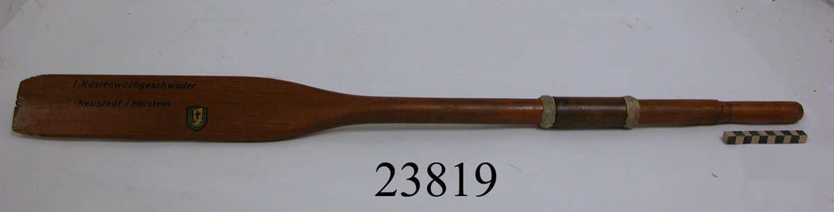 Paddel av trä. På skaftet är lindat en 80 mm bred remsa av läder. På vardera sida därom ett flätat band av vitt garn. På bladet svart text: 1. Küstenwachgeschwadre Neustadt/Holstein samt en liten målad vapensköld.