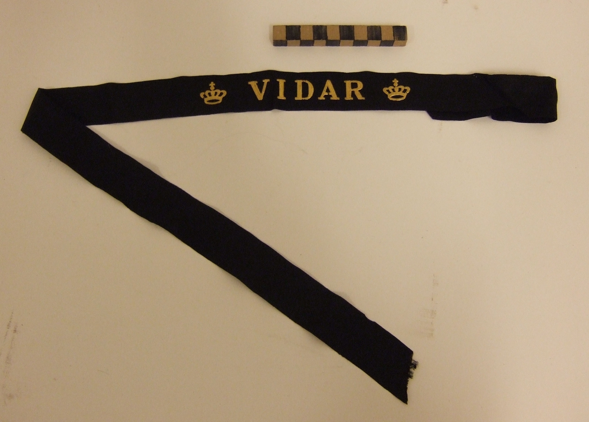 Mössband till rundmössa för manskap vid flottan.  Av svart rips med namn "VIDAR" i guldbokstäver, flankerat av två kungl. kronor i guld. 7st olika mössband i samma förpackning. Alla från samma givare. Inventariebilagan ligger under MM 24615.