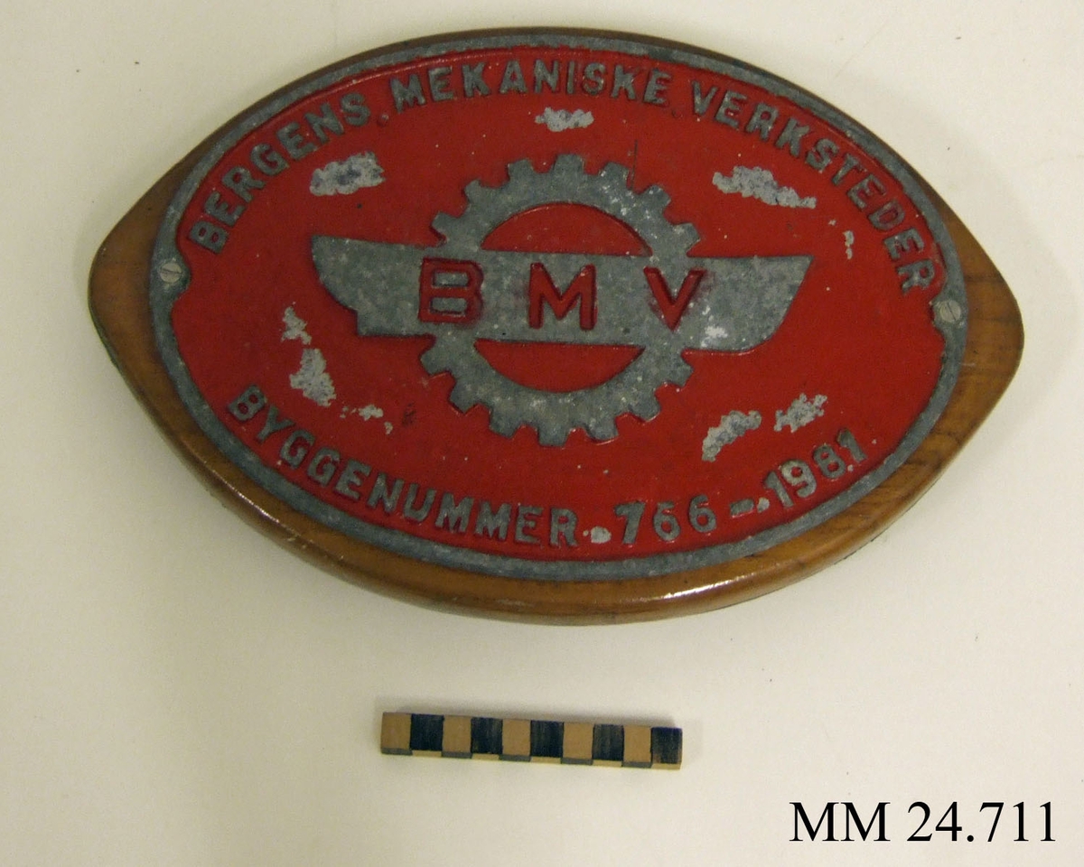 Nybyggnadsskylt till patrullbåten Spejaren. Oval, rödlackerad skylt av metall, med text i relief: "Bergens Mekaniske Verksteder, Byggenummer 766-1981" samt logotype föreställande kugghjul och skeppsiluett. Skylten skruvad fast i ovalt, fenissat trästycke.