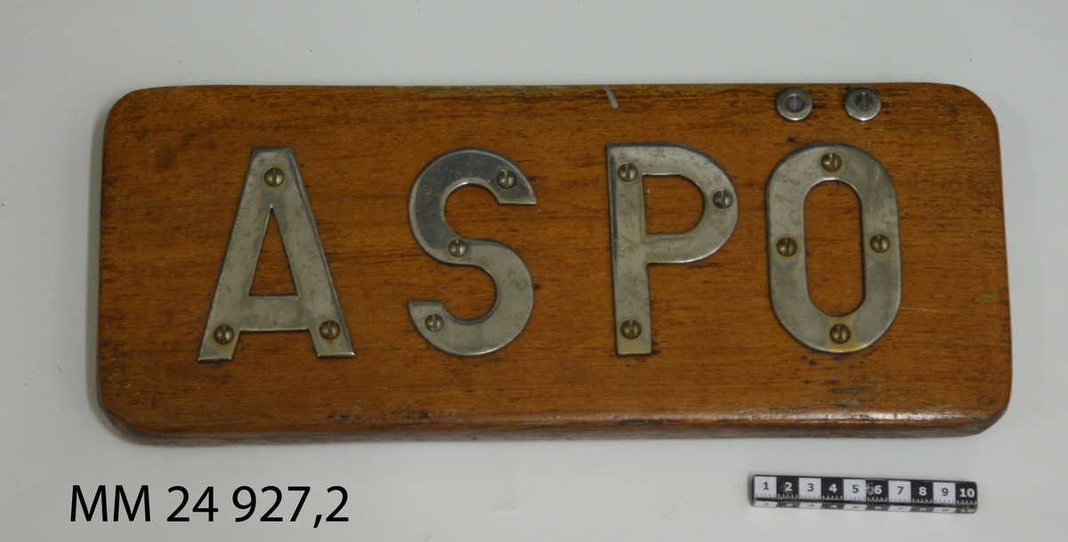 Namnbräda av fernissad teak. Bokstäver av stål fastskruvade från framsidan, bildar namnet: "Aspö".
