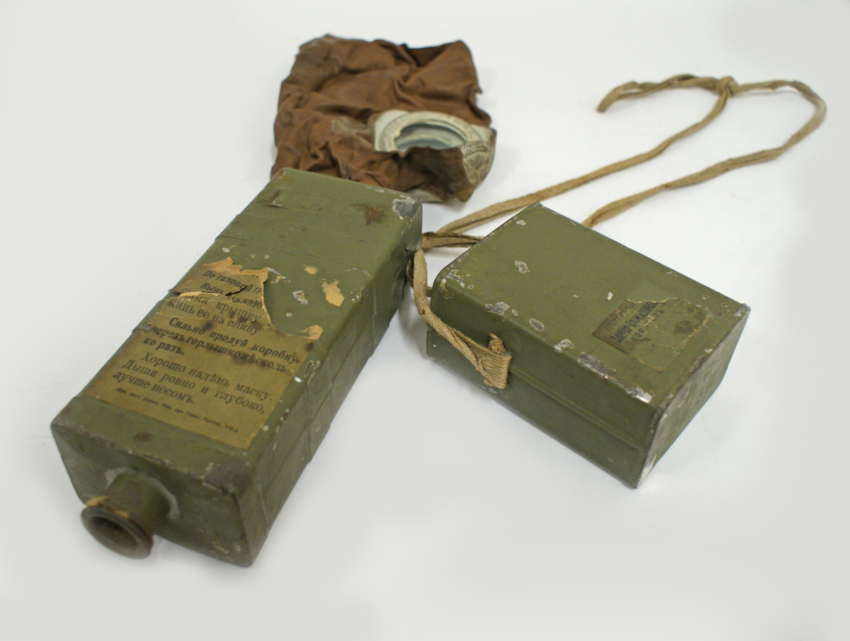 Gasmask, Zelinsky Kummant modell 16 kallad "Moscow". Rektangulär låda av plåt, grönmålad. Avtagbart lock som rymmer masken. Masken är av brunt gummi med runda titthål av glas. Gasmasken är fäst i pipa i filterlådan. Filtret är ett kolfilter med en linning av bomull som hindrar inandning av kolet. Pipa i filterlådans botten. Locket och filterlådan sitter sammen med textilsnöre som även gör att gasmasken kan hängas runt halsen. Diverse pappersettiketter med rysk text sitter på filterbehållaren: "CEPIR 296" mm.