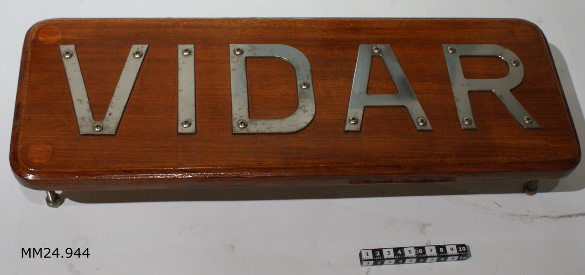 Namnbräda i fernissad teak med profilerade kanet. På framsidan bokstäverna "VIDAR" i vit metall. På baksidan sticker skruvar ut i varje hörn.