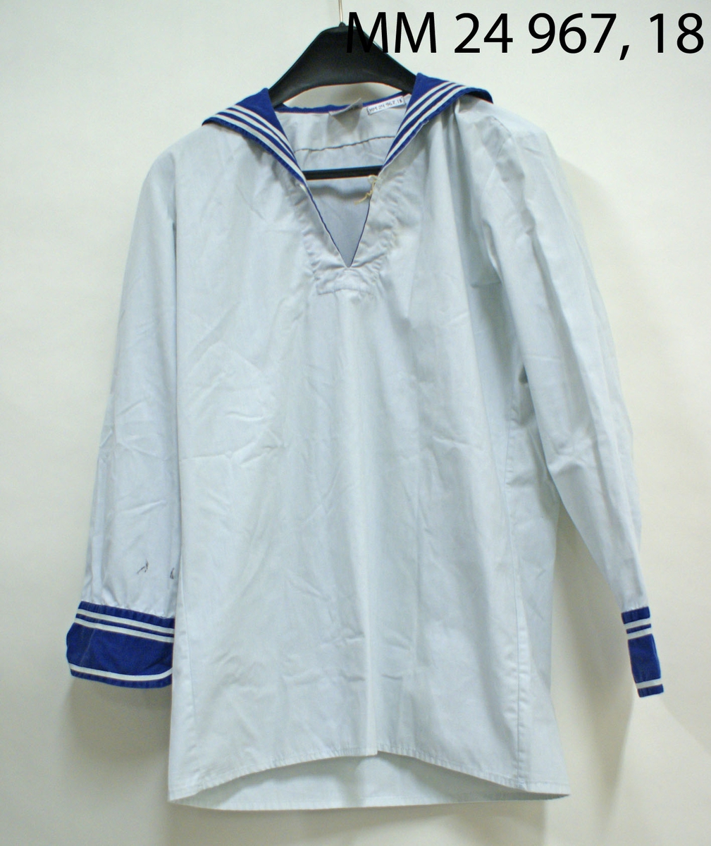 Skjorta blåkrage modell 1948. Vit skjorta av bomull och polyester. V-formad halsöppning som övergår i blåkrage. Blåkragen är fyrkantig baktill och har tre vita band påsydda längs kanten. Knapp framtill vid v-ringningen. Blåa ärmslut med vita band påsydda. Vitt lapp i nacken med tvättråd, M-nummer, kronstämpel samt årtal 1985 samt storlek 38, "Bolgo ab, tillverkad i Portugal".
