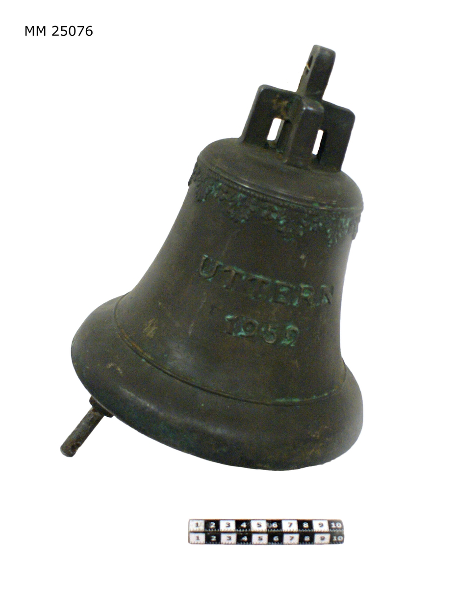 Skeppsklocka av brons märkt i relief: "Uttern 1959". Ornamentslinga i relief längs klockans topp. Kläpp av stål med kula av brons.