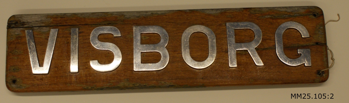 Namnbärda i fernissat trä med profilerade kanter. På brädans framside bildar bokstäver i vit metall ordet "VISBORG" i versaler. I vart och ett av de fyra hörnen är brädan genomborrad för upphängning. På baksidan stickar skruvarna som håller bokstäverna på plats fram.