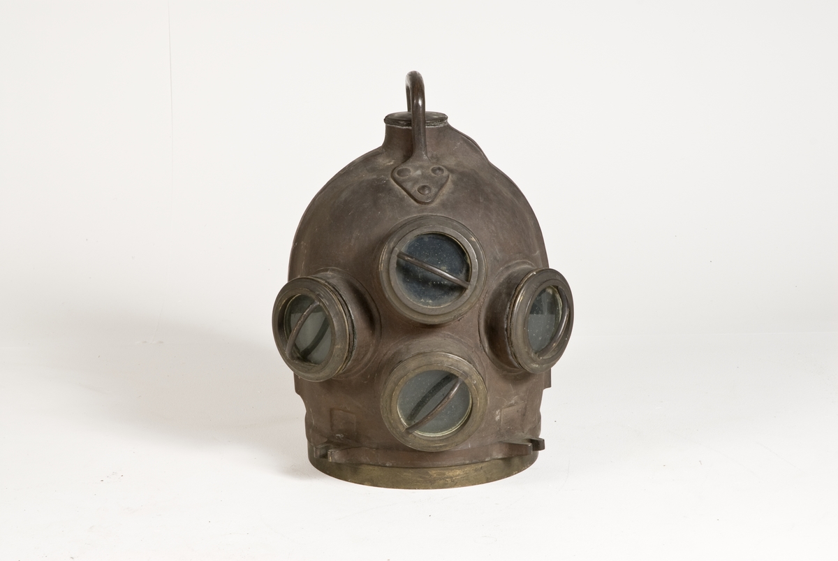 Dykarhjälm, modifierad Fahnehjelm från ca 1840-50-tal, med 4 fönster försedda med gardejärn. Upptill handtag, kort rör på baksidan för tilloppsluftslangen med backventil inuti hjälmen. Intern luftkanal från höger till vänster sida gående genom backventil med utlopp på vänstra sidan.
Från Modellkammaren.