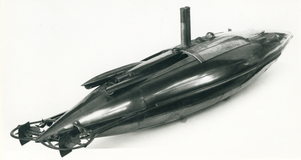 Spolformad. Stålplåt. Slingerköl, 2 propellrar av metall, 2 st höjd- och sidoroder parallellkopplade med gardjärn. 2 st höj- och sänkbara periskop av mässing. 4 st rännor för torpeder, därav 2 med luckor framtill, dels 2 för magasinering. Kort cylinder, rörlig i ränna midskepps akterut. Torpedrännorna täckas av långa stållock. Tillhörande 4 st torpeder av massiv mässing. Eneroths förslag till ubåt 1897. UndervattenstorpedFöremålets form: Spolformad.