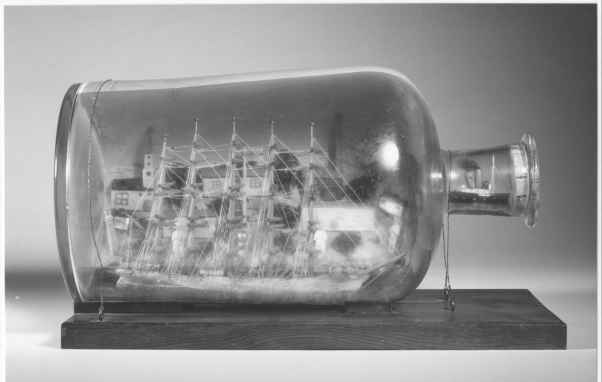 Flaskskepp med femmastad fullriggare. Seglen är beslagna och i flaskans hals är en liten bogserbåt behjälplig. I bakgrunden är en bebyggd strand. Tillverkaren är okänd.