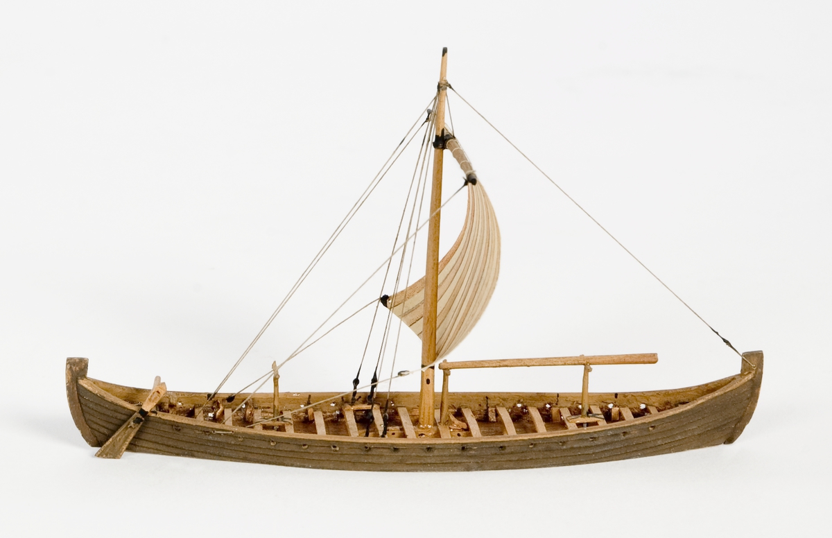 Fartygsmodell av trä. Vikingaskepp på vattenlinje. Sexton roddbänkar, styråra på styrbords sida. Riggad med en mast och ett röd-och vitrandigt segel av papper. Tältspira upplagd på bockar för om masten. Mörkbrun färg.