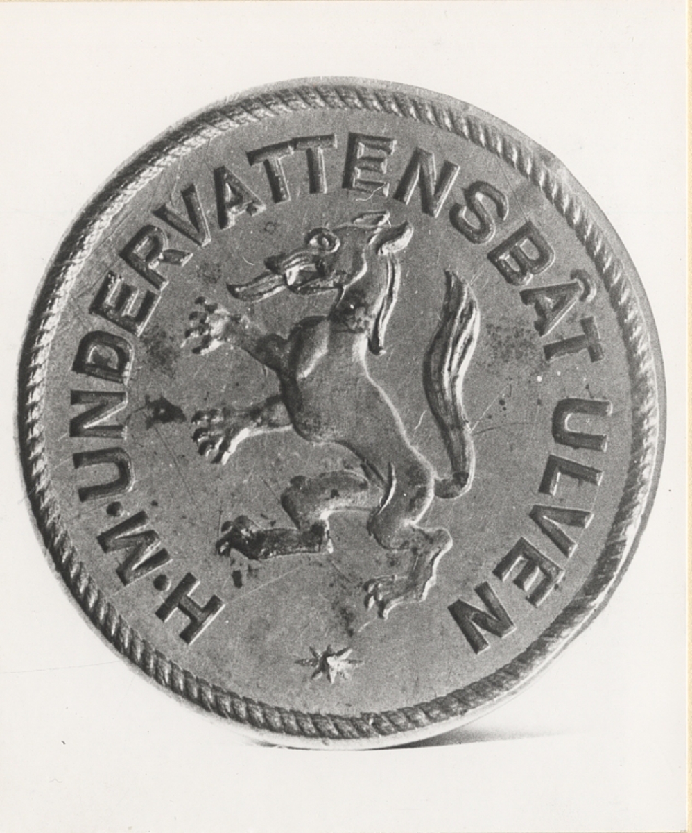 Tillhört U-båten ULVEN. Svarvat handtag av trä, svartmålat, avbrutet och lagat. Stans av mässing med bild av ulv och text enligt nedan.