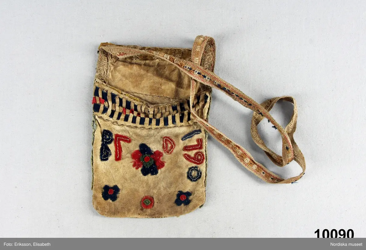Rektangulär påse av vitt skinn med enkla blomfigurer samt initialer och årtal B F D 1790 av blå, röda och gröna vadmalsremsor applicerade med efterstygn av vitt lingarn,. Kanten prydd med bård av en blå och en röd yllerema trädda genom uppskurna slitsar i skinnet, i kanten fragment av gult ylle.
Mycket slitet linneband med upphämtamönster i gult, rosa och blått ullgarn fastsytt som axelband.
Insamlat av Nils Månsson Mandelgren vid mitten av 1800-talet.
/Berit Eldvik 2010-02-19