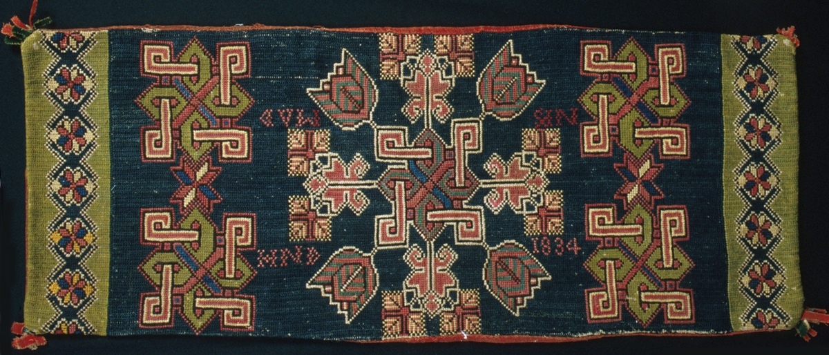 Dyna/bänklängd broderad i tvistsöm och korsstygn. En valknut på mitten med fyra tulpaner samt blommor och blad. Ytterligare fyra valknutar och två stjärnor i det mörka mittpartiet. En rosbård längst ut i kortsidorna. Mörkblå botten i mittpartiet och ljusgrön botten i bården. Mönster i två nyanser grönt, rött, gult, naturvitt, blått och brunt. Tofsar av rött, grönt och blått siden, bomulls- och ylletyg i hörnen. Märkning i korsstygn i rött: "HND  1834  NPS  MAD". Sydd på oblekt linneväv i tuskaft med dubbelt 1-trådigt ullgarn. Botten och de flesta mönster är sydda i tvistsöm, enstaka stygn, diagonala linjer och konturer är sydda i korsstygn. Baksida av rött ylletyg vävt i tuskaft, skarvad och trasig. Öppning mitt på bakstycket. Påsydd tyglapp på baksidan med texten: "A. N° 95 a  Landstinget  Torna h-d".