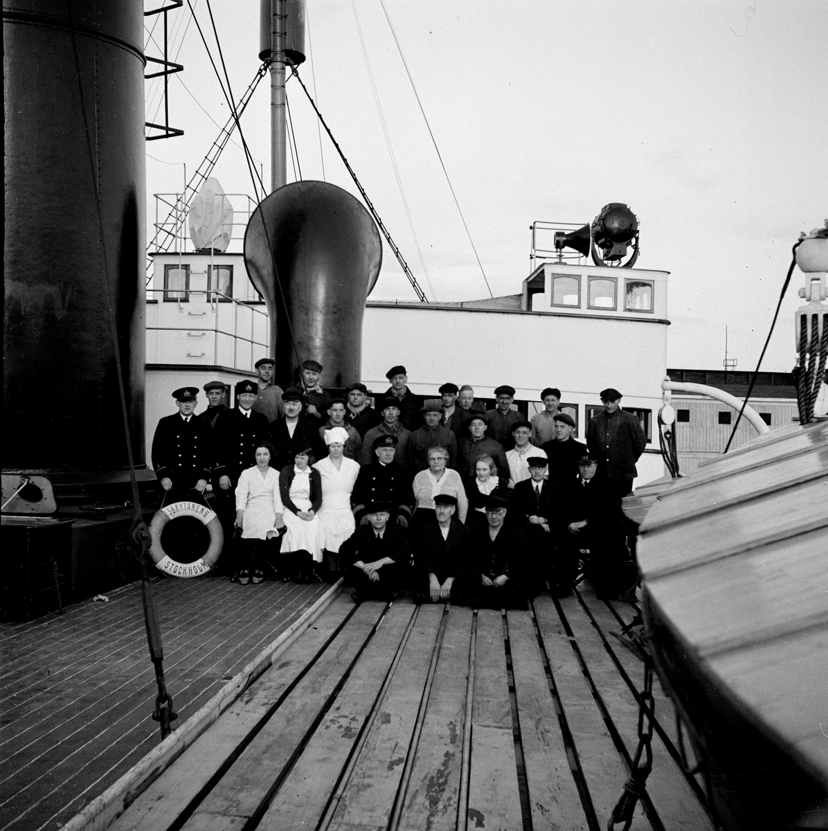 från Fotobeskrivning:] "ISBRYTAREN II:s besättning på båtdäck. Sittande på däck: maskinist Sven Lindqvist, maskinist Linderlöv, maskinist Tjernberg. Sittande på stolar: okänd, okänd, okänd, kapten Georg Hilding, restauratris Anna Andersson, okänd, övermaskinist Bror Erik Nilsson, styrman Sven Noedqvist [sic]. Stående: telegrafist Eric Hedström, oidentifierad smörjare, styrman Evert Brolin, okänd, 2:a maskinist Björkström, övriga okända."