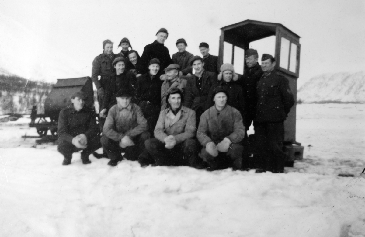 Norske anleggsarbeidere (?) og et par tyske soldater poserer foran et lite hus/bu og en slags tippvogn.