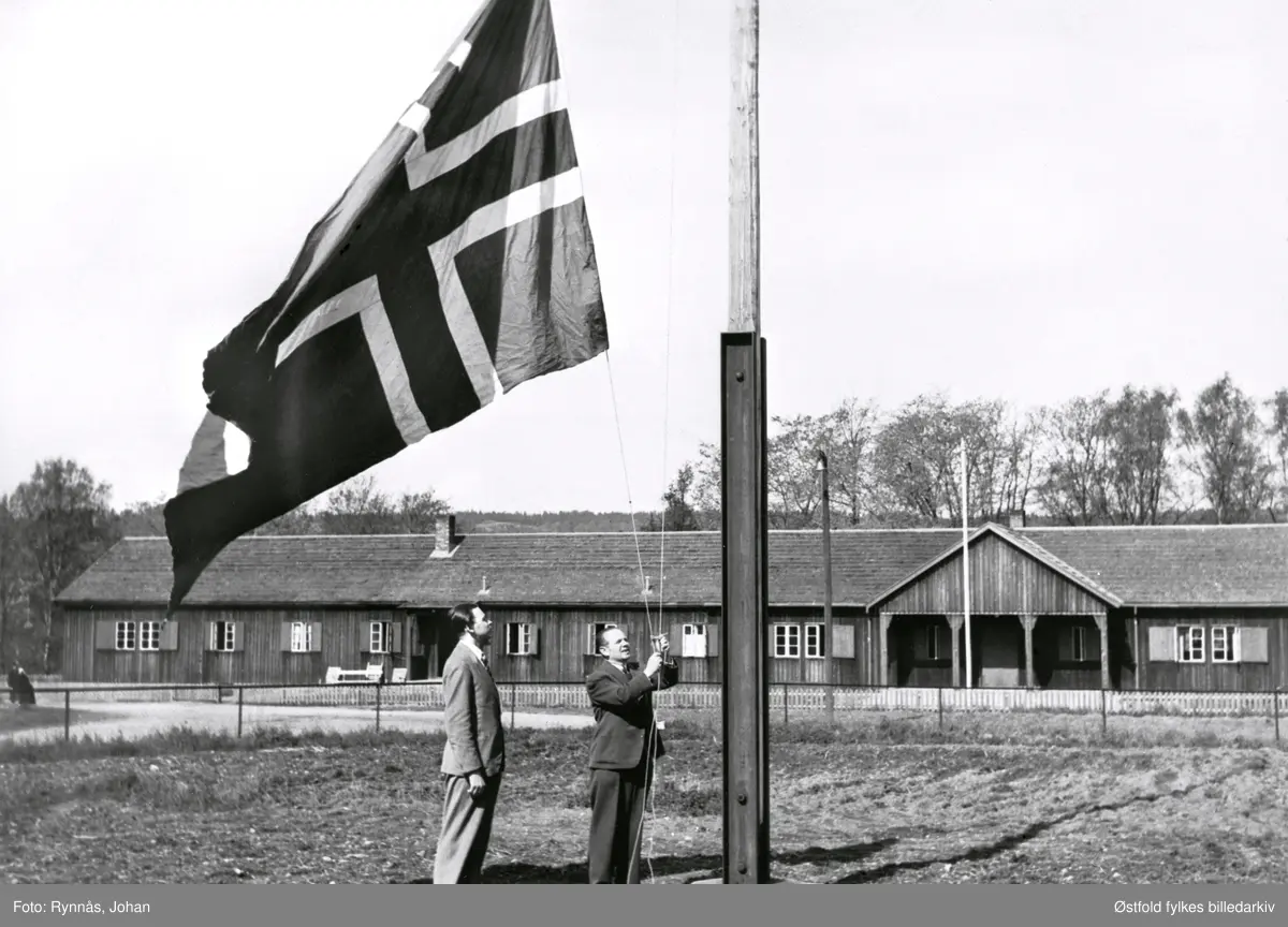 Rådmann Alf Herland og sekretær Arne Kinander i Moss kommune heiser det norske flagg under frigjøringen 8. mai 1945 i Moss.

"Flagges heisen på eiendommen Thorneløkka, hvor Moss kommune hadde holdt til fra 1921. De kjøpte bygningen Breidablikk på Thorneløkka. I 1974 ble et nytt kommunehus oppført og Breidablikk, med andre bygninger på tomta ble revet. Det nye kommunehuset som fortsatt er i bruk i dag ble designet av arkitekt Jan Jansen. 

I bakgrunnen ser vi rester etter tysk okkupasjon. En såkalt «Todt-brakke». Tyskerne hadde flere hovedkvarter rundt i Moss, og slike brakker ble satt opp til husvære for de tyske soldatene. Okkupantenes «Organisation Todt Einsatzgruppe Wiking», sto bak slike brakker. De ble satt opp over hele landet og det gikk svært raskt å bygge dem. Brakkene ble brukt som fangeleir og lager ved siden av husrom for soldatene. Brakkene hadde navnet «Tyskerbrakker» på folkemunne. Etter krigen ble brakkene i Moss tatt i bruk av lag og foreninger, samt noen av dem omgjort til leiligheter. Blant annet den som er avbildet. Brakka ble revet i 1968.

Arne Kinander var kinosjef i mange år etter han var ferdig i sekretærstillingen. Han var blant annet sjefen i sekreteriatet for den store Mosseutstillingen i 1937.  I 1970 hadde han igjen en stor rolle i byens 200-års jubileum og på sine eldre dager var han sekretær for kommunens bokkommite. De arbeidet med bokserien «Moss bys historie »

Rådmann Alf Herland, satt som rådmann i Moss kommune både før og etter 2. verdenskrig. Herland var svært godt kjent i Moss, og i 1970 ga han ut boka "Gatenavnene i Moss".
Forfattet av Arve Tomt Gundersen/Mossehistorien.

http://mossehistorien.blogspot.no/