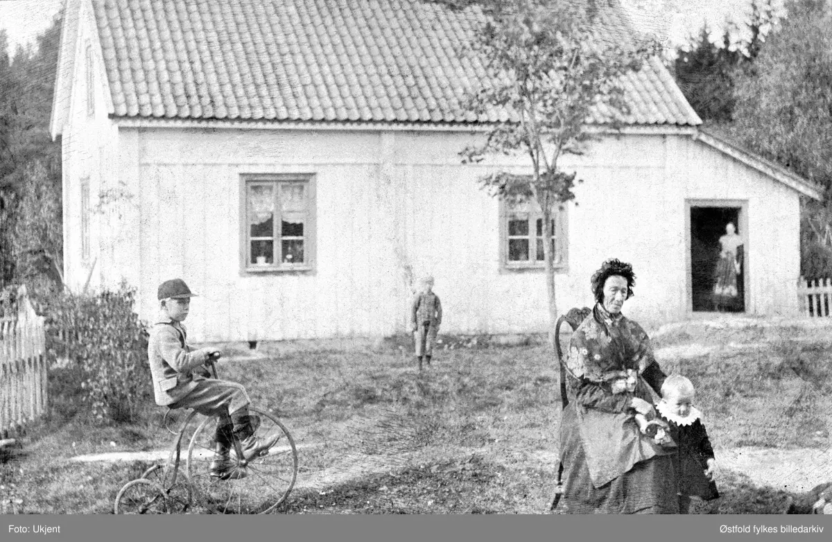 Barn og bestemor på tunet, Haga gård ca. 1892. Gutten på tre-hjulssykkelen (velociped) eller veltepetter er Eivind Haga f. 1884 Damen på stolen er hans bestemor Petrine Haga, F. Hjelmark. Jente med hvit krage er Petrine Svenneby f. 1891, F. Høie.