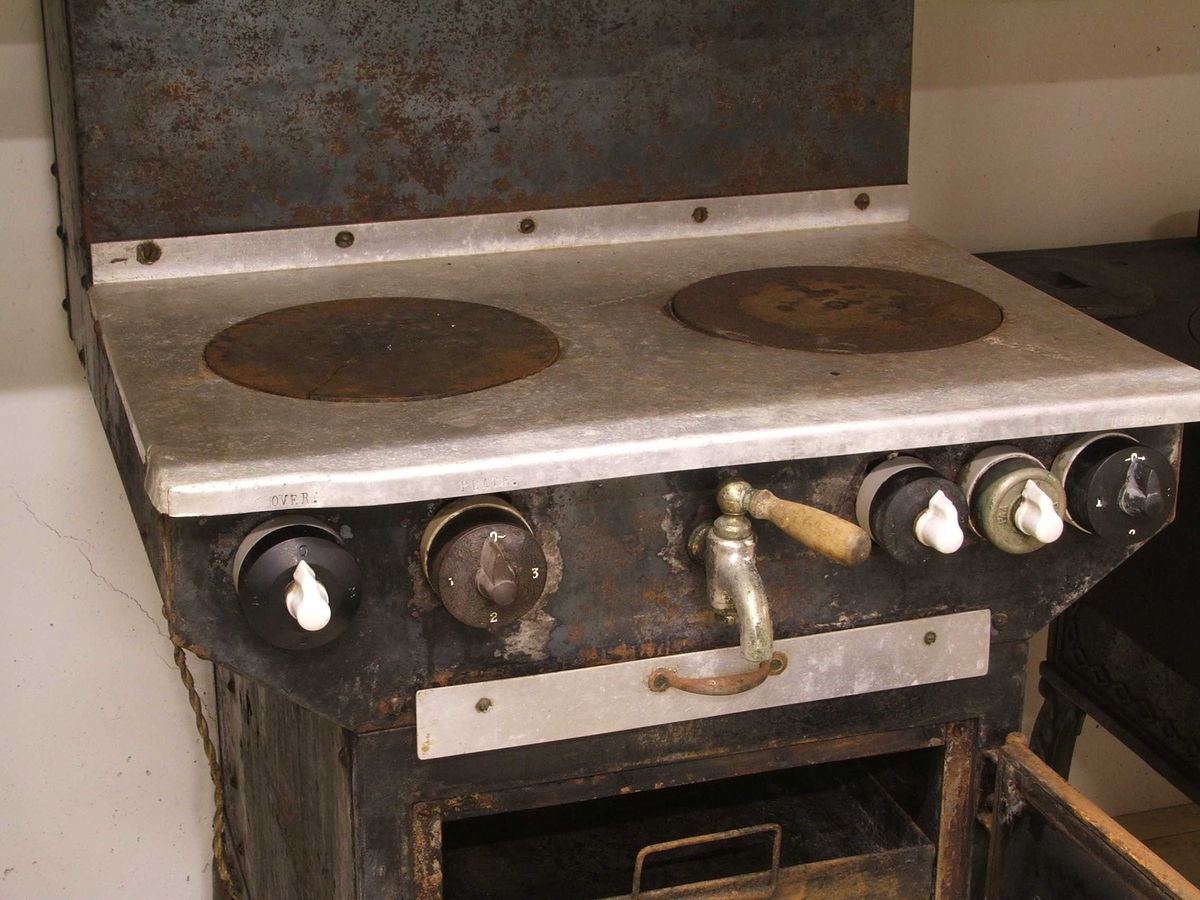 Flølo komfyren. Elektrisk komfyr. Med tank for varmt vatten (oppå bak), og kran for tapping av det varme vatnet framme. To kokeplater oppå. Fem runde brytarar framme i front. Ei uttrekkbar plate under. Omnsdør til å opne inn til steikjeomnen under der att. To plater med handtak inne i steikjeomnen. Komfyren står på fire bein (av jarn).