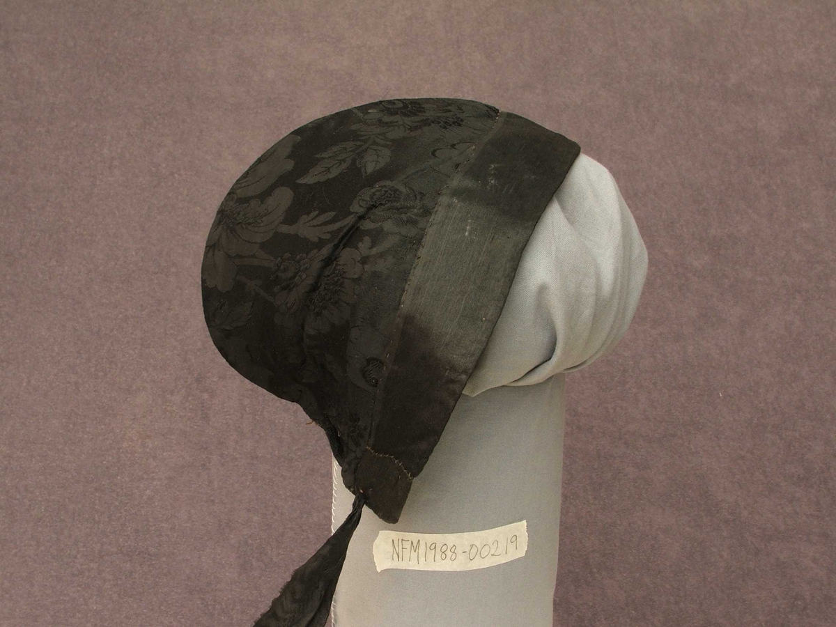 Svart "hue" til kvinne. Konehue. Typen er tett inntil hovudet. Med 4 cm kant fremst i glatt stoff, og med resten av utsida i svart stoff med innvove blomemønster.Lange band bak i nakken til knyting. Bak i nakken ein om lag 11 cm høg splitt, øvst i splitten er bandet trædd inn og i to hol på kvar side. Det er altså eitt band som kan snurpast ihop her. Fôra med lys gult og kvitt bomullsstoff.