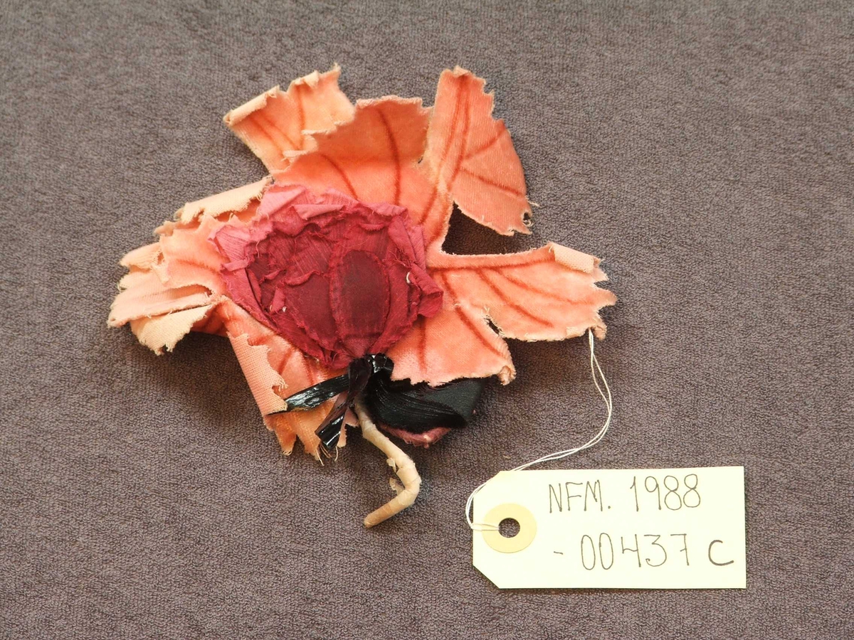 Seks ulike stoffblomar til pynt på drakt eller hatt. a: Bukett - orange/gul. b: Bukett - kvit/grøn. c: Rosett - svart/burgunder/rosa. d: Bukett - grøn/lilla. e: Bukett - grøn/lilla. f: grein - lys rosa. 