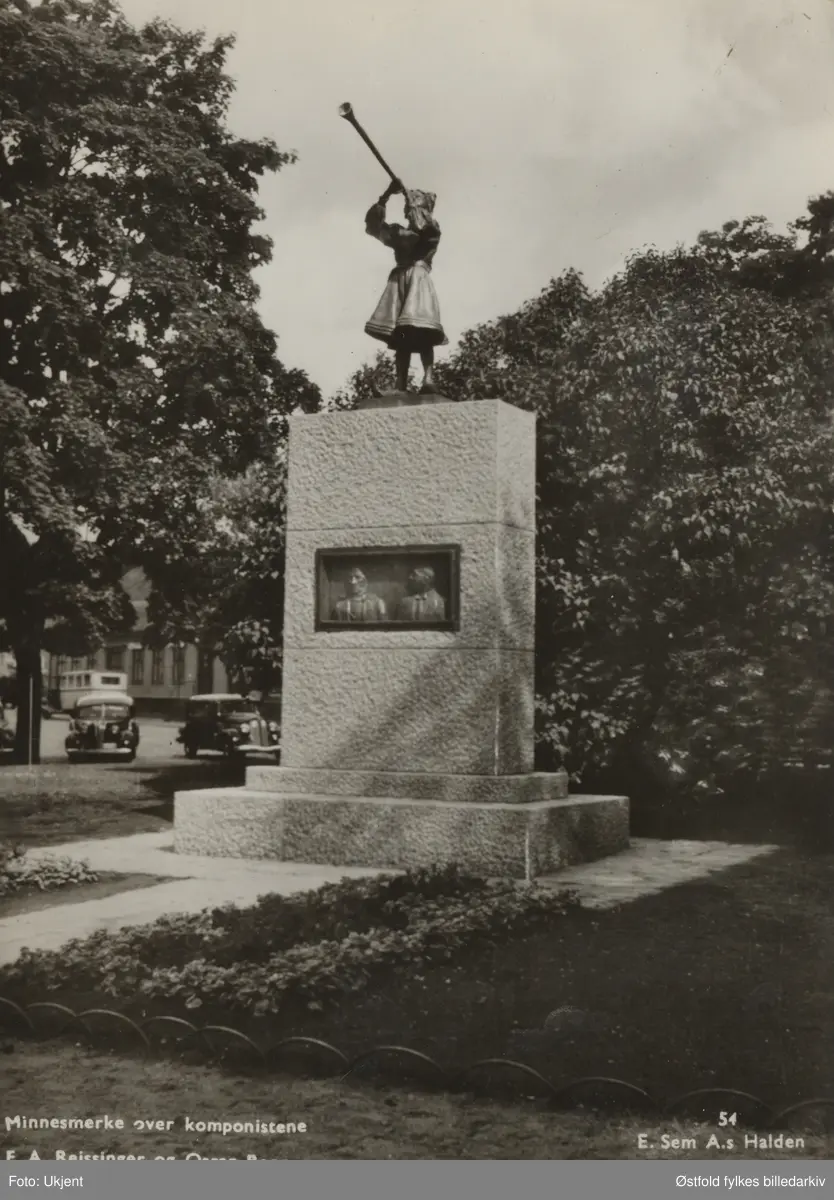 Minnestøtte over komponistene F. A. Reissiger (1809-1863) og Oskar Borg (1851-1930) i Busterudparken i Halden.. Innfelte portretter av kunstnerne ses på støtten og på toppen stårei jenteog blåser i et horn. Bak støtten ses noen biler og en liten buss. Bilen rett foran bussen er en Hudson eller Terraplane årsmodell 1936-37. Postkort.