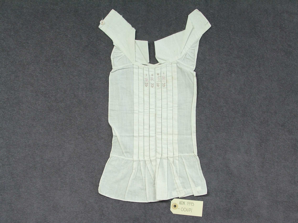 Skjortebryst til å ha framme på brystet. Til mann. Forseggjort saum. Krage til å knappe bak med knapp og knapphol. Eit knapphol på kvar side av kragen. Fire pyntefaldar på kvar side av midten framme på brystet. Det er ein slags netting langs midten og holsaum mellom kvar fald. Sydd på omlag 10 cm kant nedst utan pynt. Brodert fire små hjarte i rosa og brune krossting under fire bokstavar og ståande firkantar over. Alt er kanta.