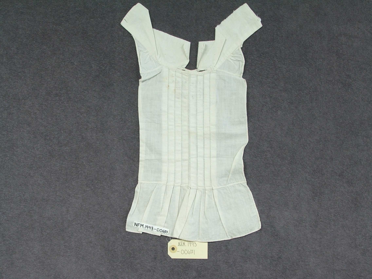 Skjortebryst til å ha framme på brystet. Til mann. Forseggjort saum. Krage til å knappe bak med knapp og knapphol. Eit knapphol på kvar side av kragen. Fire pyntefaldar på kvar side av midten framme på brystet. Det er ein slags netting langs midten og holsaum mellom kvar fald. Sydd på omlag 10 cm kant nedst utan pynt. Brodert fire små hjarte i rosa og brune krossting under fire bokstavar og ståande firkantar over. Alt er kanta.