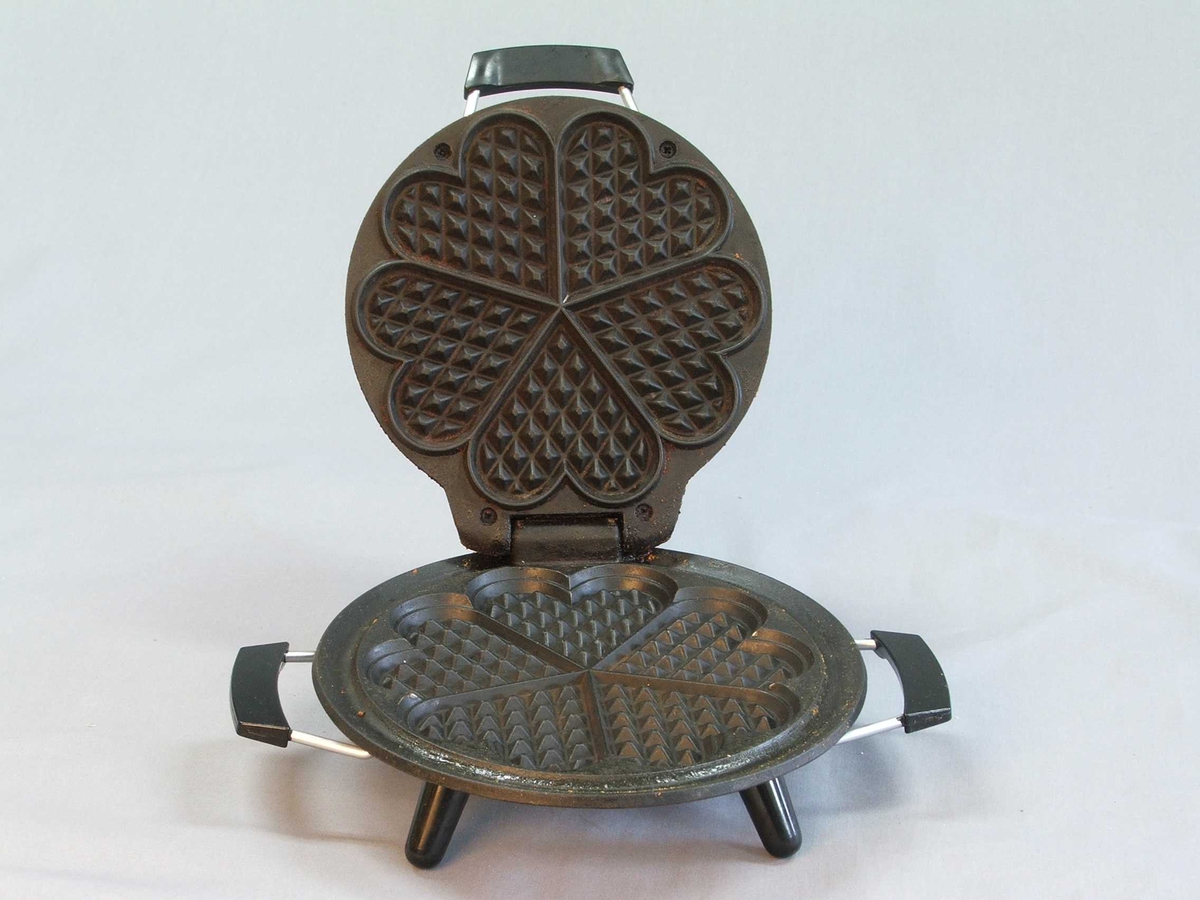 Eit vaffeljarn i original øsjke, isopor i øskja som jarnet står på. Det er ikkje ledning. Jarnet: Rund form. Loket opnast med handtak midt framme. Loket er hengsla fast til underdelen. Loket kan stå oppe av seg sjølv. Utsida av jarnet: oppå og under er det blankt metall, svart handtak på lok og to på kvar side av understellet. Inni er jarne svart, støypejarn? Vaffelhjarte-formar inni jarnet både på lok og botn = fem små hjarte som dannar ein sirkel. Fire svarte bein på undersida som jarnet står på.
Øskja er grøn med ein gul vaffel på oppsida og eit kvitt hjarte på sida. Øskja kan du opne framme. Den er kvit inni.