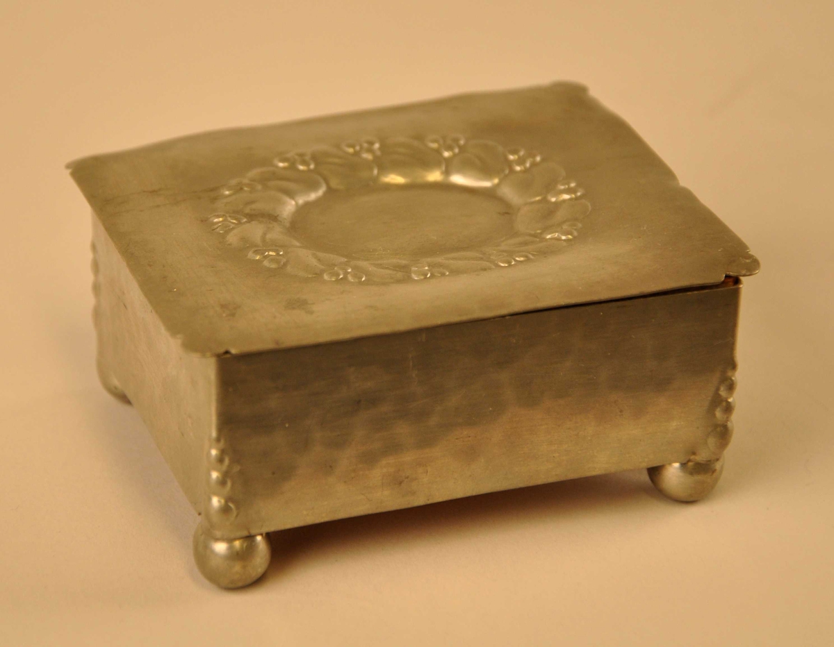 Oval  jugendstilornamentikk på loket i form av stiliseerte blad med bær. Bæra går att i hjørnedekorensom endar som kuleforma føter.