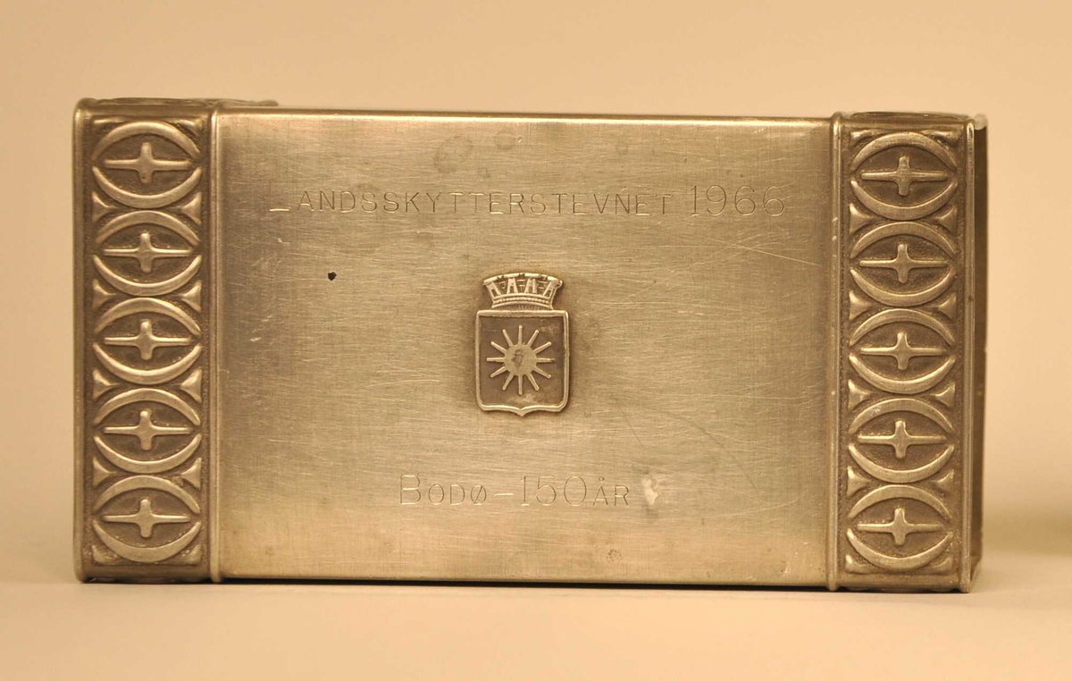 Dekorbord med tettståande, opne elipser med ein kross inni. Mønsteret går over 3 sider fyrstikkøskjeholdaren i kvar ende. På oppsida er byvåpenet til Bodø festa på  - eit skjold med ei sol med 12 strålar og ein krunelignande form samansett av 4 tårn. 