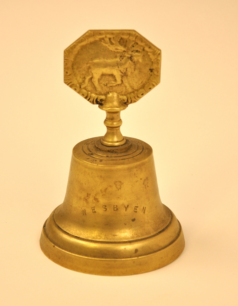 Bordklokke i messing med handtak. Klangdelen har klassisk klokkeform. Handtaket er pynta med ei åttekanta plate med  eit reinsdyrrelief på  og skrudd saman med underdelen. På innsida heng pendelen i skrutaket etter ein krok og har ei støypt messingkule som slag